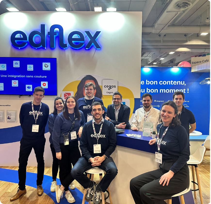Groupe de neuf personnes souriantes portant des badges et des vêtements edflex, posant dans un stand d'exposition moderne avec un grand logo edflex lumineux et un écran affichant des services numériques.